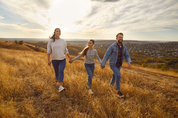 Happy smiling young family with child girl holding hands walking in the field enjoying nature at sunset. Mother, father and their daughter spending time outdoors. Family leisure concept. - obrazy, fototapety, plakaty