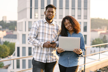 Portrait of young adult man and woman using laptop while standing outdoors office building. Two...