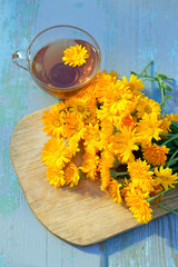 Fresh yellow-orange calendula flowers and glass cup with herbal tea on rustic wooden table. Healing infusion with medicinal plant calendula. useful anti-inflammatory, antiphlogistic drink. top view.