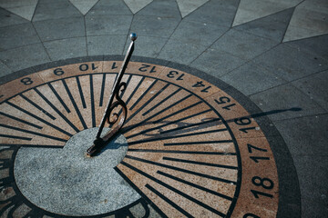 A sundial on the sidewalk of the city square. The arrow casts a shadow. The clock shows the time