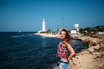 A girl is stands on the edge of a cliff of a sea cliff against the background of a white lighthouse. The girl closed her eyes in pleasure. The girl is wearing denim shorts and a red plaid shirt