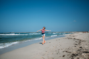 A girl in denim shorts and a red plaid shirt runs along the ocean on the snow-white sand. The woman...