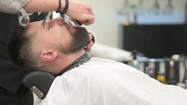 A hairdresser forms the outline of a beard with wireless trimmer in a barbershop.