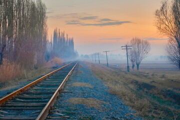 Fototapeta premium A railroad disappearing into the distance amidst fields in rural scenery, accompanied by old wooden electrical poles running alongside, against the backdrop of a sunset sky and distant light mist.