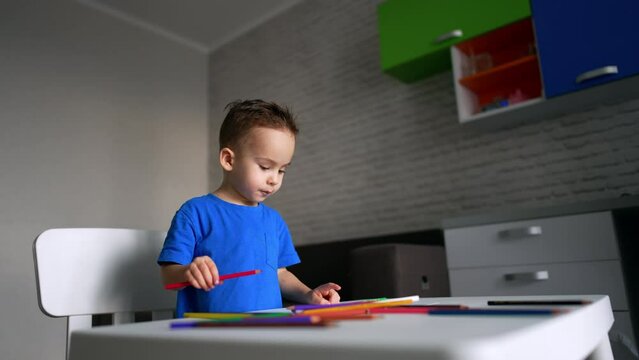 Dark-haired Caucasian toddler boy in blue t-shirt stands at his desk. Cute baby drawing with a red pencil. Low angle view.