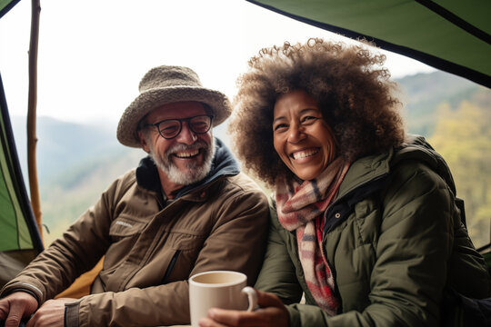 
Happy senior biracial couple sitting in a tent and drinking coffee in the mountains