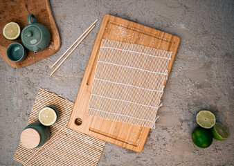 Wooden cutting board, bamboo sushi mat, chopsticks, fresh lime wedges, ceramic green teacup on a...