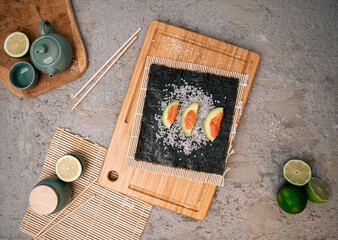 Nori seaweed with rice grains, sliced avocado with salmon on a wooden cutting board, chopsticks,...