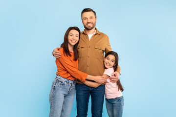 Happy European Family Of Three Standing Embracing Over Blue Backdrop