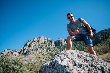 A male tourist on the background of high stone mountains. The man stands with one foot on a stone and smiles. The man is wearing sunglasses and a travel backpack