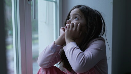 Sad pensive little girl feeling solitude by apartment window staring at view with thoughtful gaze....