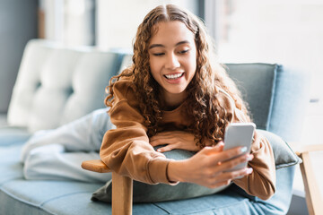 Cheerful teenager girl smiles using phone for online activities indoor