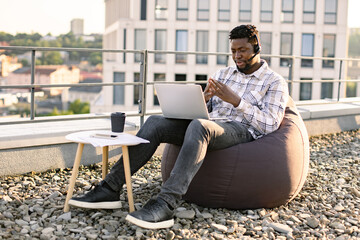 Relaxed African American man conducting web conference via laptop and headset while staying on flat...