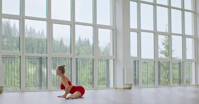 Young woman practicing yoga split pose in spacious studio. Side view of graceful female wearing red activewear stretching body in leg-split, against mountain landscape, motion. Fitness concept.
