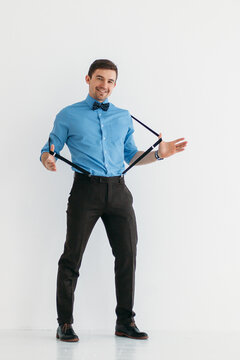 A man in a blue shirt, classic trousers, suspenders and a bow tie against a white wall in the studio. The man pulls back his suspenders with both hands and smiles  broadly