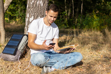 A man is sitting in a park near a tree and charging his phone from a solar panel