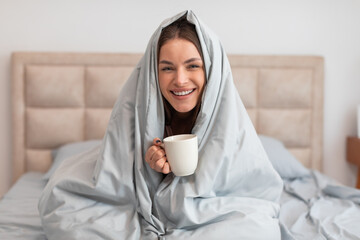 Woman wrapped in duvet holding coffee mug, enjoying morning