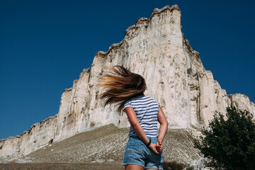 A woman in the background is a white rock with a steep cliff. Scorched grass from the summer heat. The woman is wearing denim shorts and a vest. The woman jumped up, her hair flying in the wind