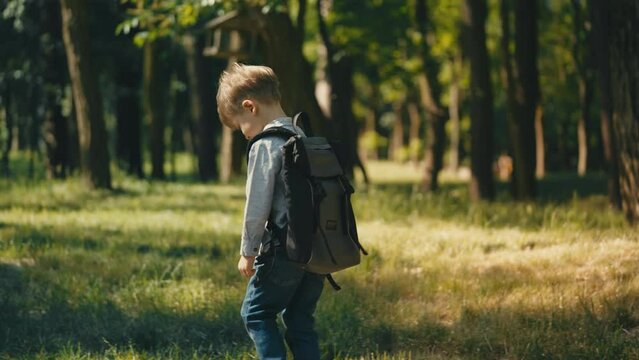 Shy little boy with school bag walks in park alone, enjoys rest after classes