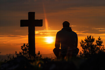 Silhouette of praying man at hilltop cross with warm glow of a sunset. Hope for salvation, request for help to heaven. Concept of faith in God
