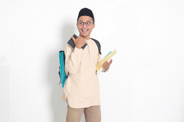 Portrait of excited student Asian muslim man in koko shirt with skullcap carrying backpack, holding...