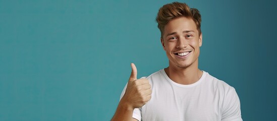 A young Caucasian man dressed in a white t-shirt confidently extends his arm upward, giving a thumbs up gesture with a happy smile on his face. - obrazy, fototapety, plakaty