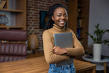 Professional woman holding closed laptop while standing in home office interior