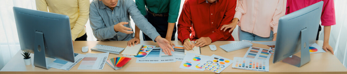 A top view cropped imaged of creative graphic team selects colors carefully from color wheel and palette on table at modern studio, surrounded by graphic documents and materials. Variegated.
