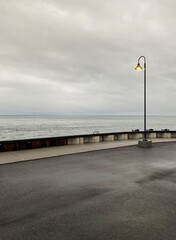 Lone lamppost at the end of a quay on a cloudy, gray day. Pier in the sea. A harbor bank and river waves in the distance on the horizon.