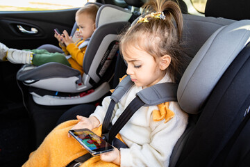 Cute siblings sitting in baby booster car seats and using mobile phones during the road trip....