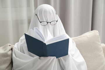 Creepy ghost. Person covered with white sheet reading book on sofa at home