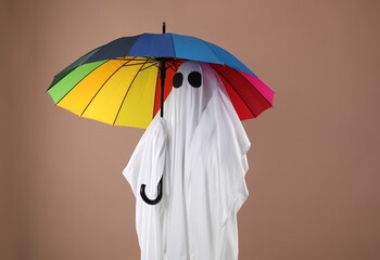 Person in ghost costume with rainbow umbrella on dark beige background - obrazy, fototapety, plakaty