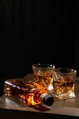 Whiskey with ice cubes in glasses and bottle on wooden crate against black background, closeup. Space for text