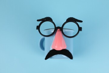 Man's face made of cup, fake mustache, nose and glasses on light blue background, top view