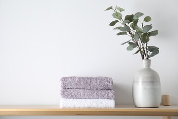 Stacked terry towels and eucalyptus branches in vase on wooden shelf near white wall, space for text