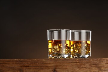 Whiskey with ice cubes in glasses on wooden table against brown background, space for text