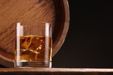 Whiskey with ice cubes in glass and barrel on wooden table against black background, space for text