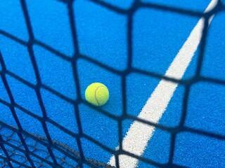 Close-up view of a line and texture of blue paddle tennis court and lonely yellow tennis ball. Artificial grass paddle court surface.