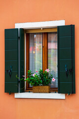 Picturesque colorful wooden old style window with green shutters and flowers on windowsill. Bright orange house on Burano island, Venice, Italy