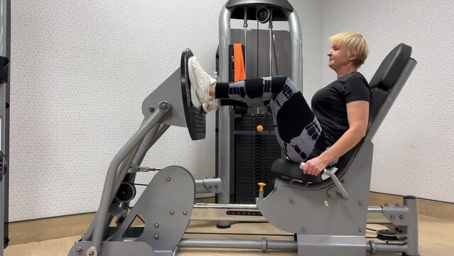 Footwork video of leg press hamstring training. Purposeful woman working on simulator in gym. Close-up video of a European secluded middle-aged woman on a leg exercise machine. A 55-year-old blonde