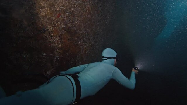 Freediver swims underwater in the sea. Man freediver floats in the underwater canyon and explores it with torch