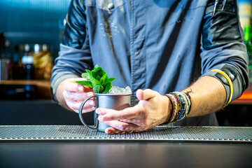 Barman prepara un mojito cocktail en la barra de un cocktail bar exclusivo vestido con una chaquetilla azul con iluminación nocturna, detalle close up