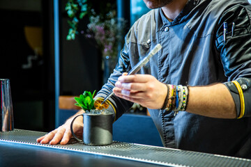 Barman prepara un mojito cocktail en la barra de un cocktail bar exclusivo vestido con una chaquetilla azul con iluminación nocturna, detalle close up