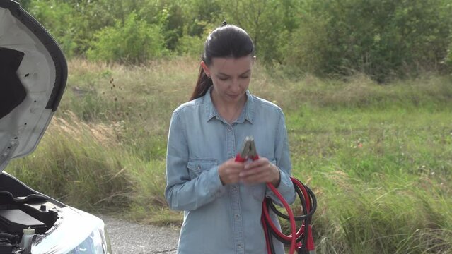 A female driver stands by the roadside next to a broken-down car, attempting unsuccessfully to jumpstart the vehicle by connecting ignition cables.