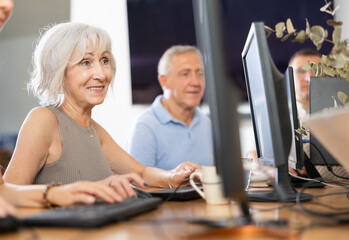 Interested positive elderly woman using computer to surf internet in library. Concept of...
