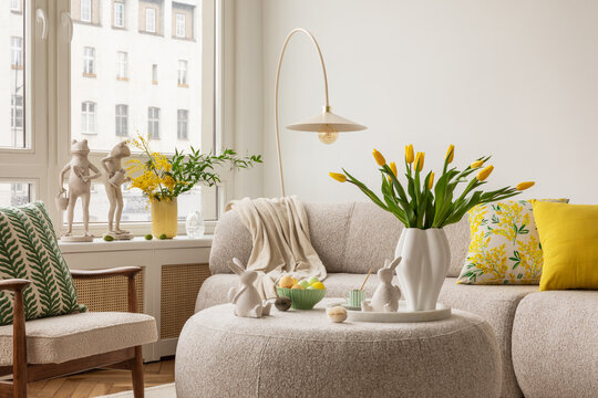 Interior design of spring living room with design sofa, furniture, vase with tulips, easter decorations, pillows and personal accessories. Home decor. Template. Easter holidays.
