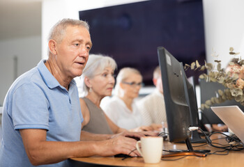 Concerned old man sitting at computer together with other attendees of IT courses