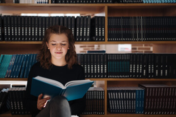 A focused teenage girl reads a blue book in a library, surrounded by shelves of literature