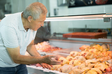 Interested elderly man looking at large assortment of fresh raw meat products in glass refrigerated display case in butchery shop - obrazy, fototapety, plakaty