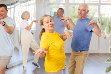 Cheerful enthusiastic senior lady enjoying active dancing paired with male partner during group training in dance studio, practicing playful twist..
